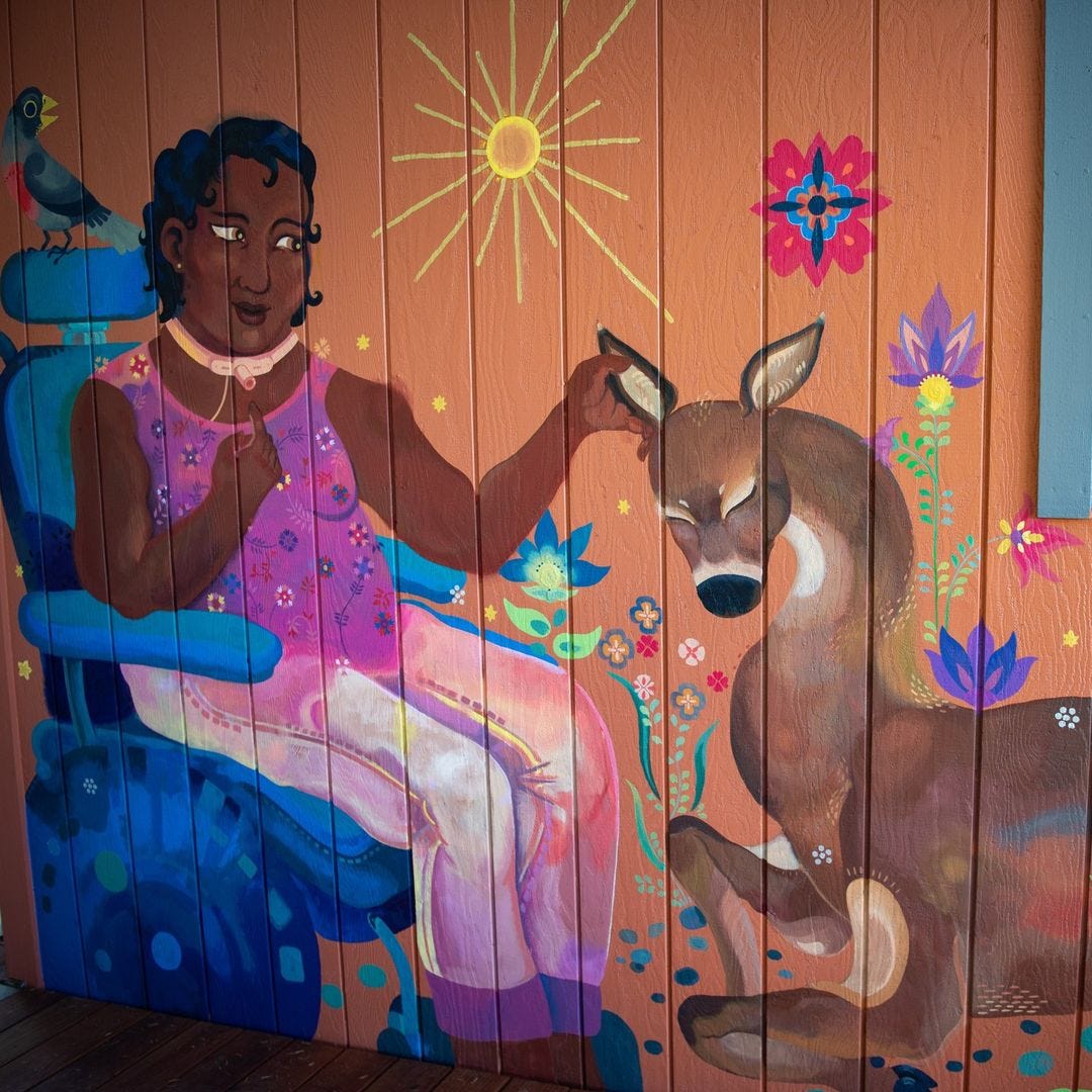 A colorful mural on the outside wall of a building shows a Black woman with a trach caressing the ear of a calm deer. A bird perches on the headrest of their powerchair. The sun beams down on blessedly blooming flowering plants.