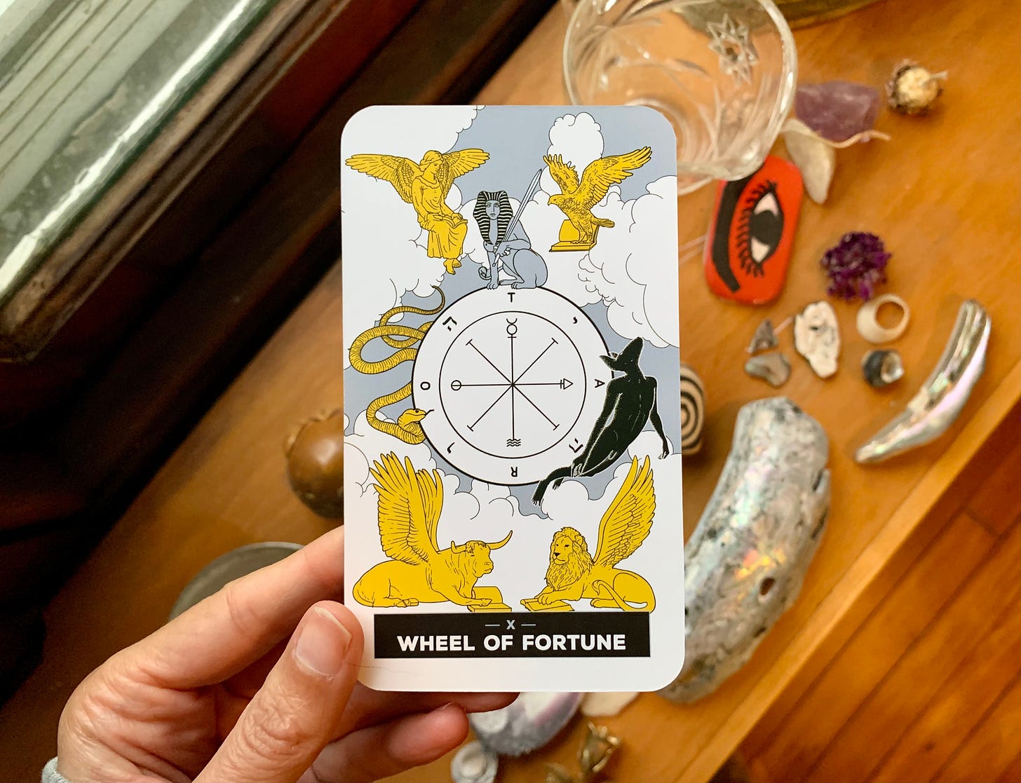 A hand is holding a tarot card, Wheel of Fortune by Xaviera Lopez for The Change Tarot. In the image, there is a wheel with a snake on one side and a demon on the other. There is a sphinx holding a sword at the top. On each corner of the card are four winged beings, a human, eagle, lion and bull. Behind the card is a wooden shelf with various objects on it including shells, fried flowers and talismans.