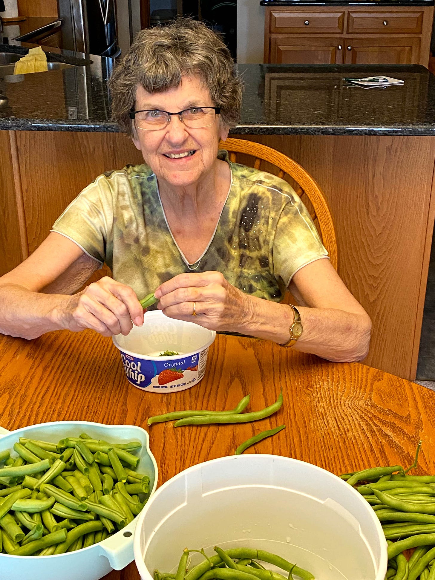 Author's mother sitting at a table, snapping a bean, and smiling.