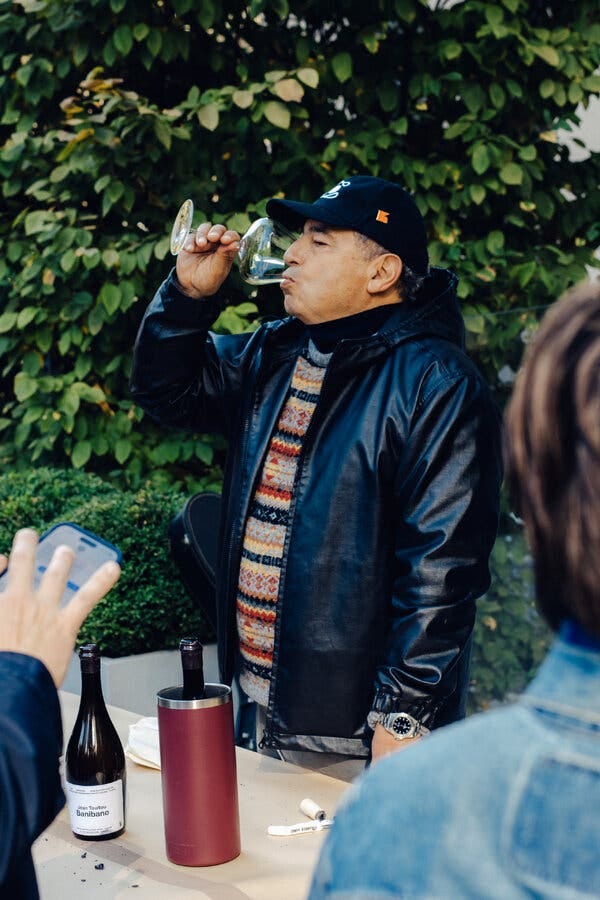 A man in a sweater and a waist-length leather jacket sips from a wine glass.