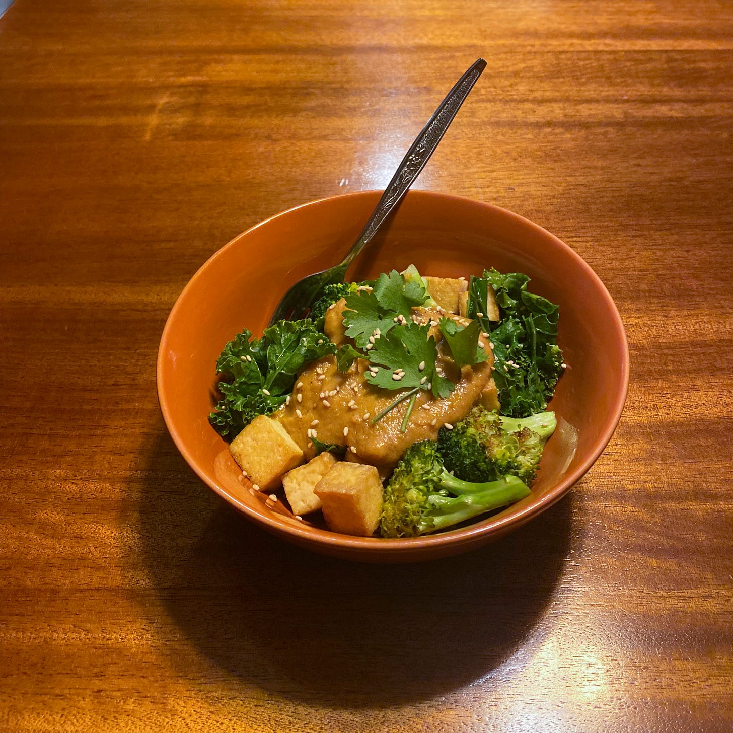An orange bowl with kale, fried cubes of tofu, and seared broccoli topped with a thick peanut sauce, cilantro, and sesame seeds. There is brown rice underneath, but it's not visible.