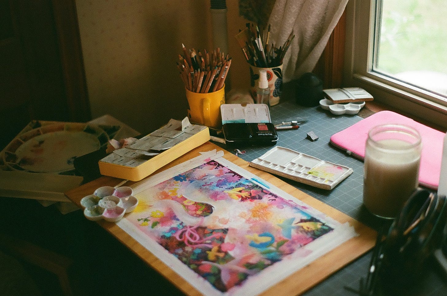 Film photo of the illustrator's studio desk. A work in progress illustration is surrounded by art supplies, including watercolor, gouache, pencils, pens, brushes, and more.