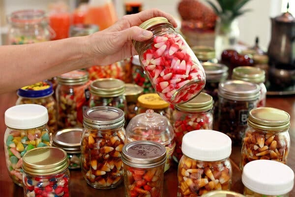 A dozen or so mason-style jars are arranged on a table. Each is filled with different flavors of candy corn. A hand is reaching in from the top to pick up one of the jars.