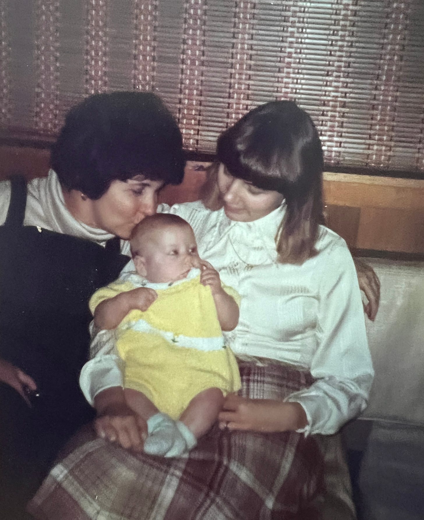 Gram kisses the head of Baby Patty, who is dressed in yellow and held in Mom's lap as Mom looks down at Patty.