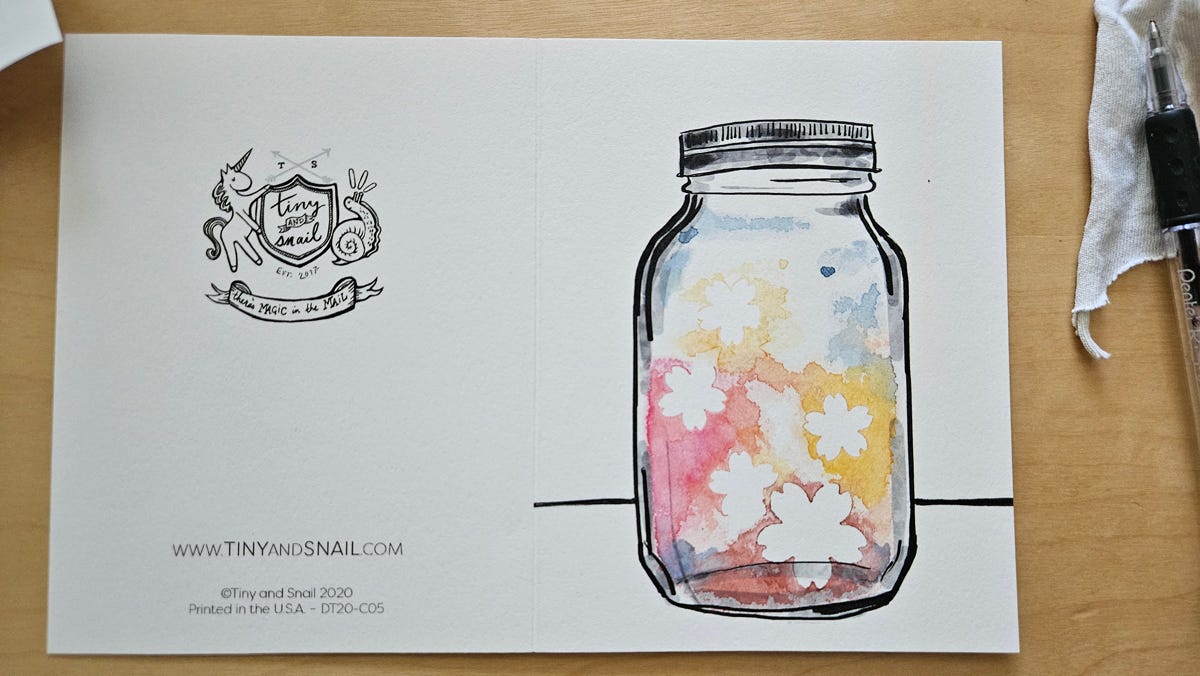 A flat view of the card, showing the logo of the Tiny and Snail company on the card back and the black mason jar line drawing painted with watercolors around white flowers previously masked by blue tape