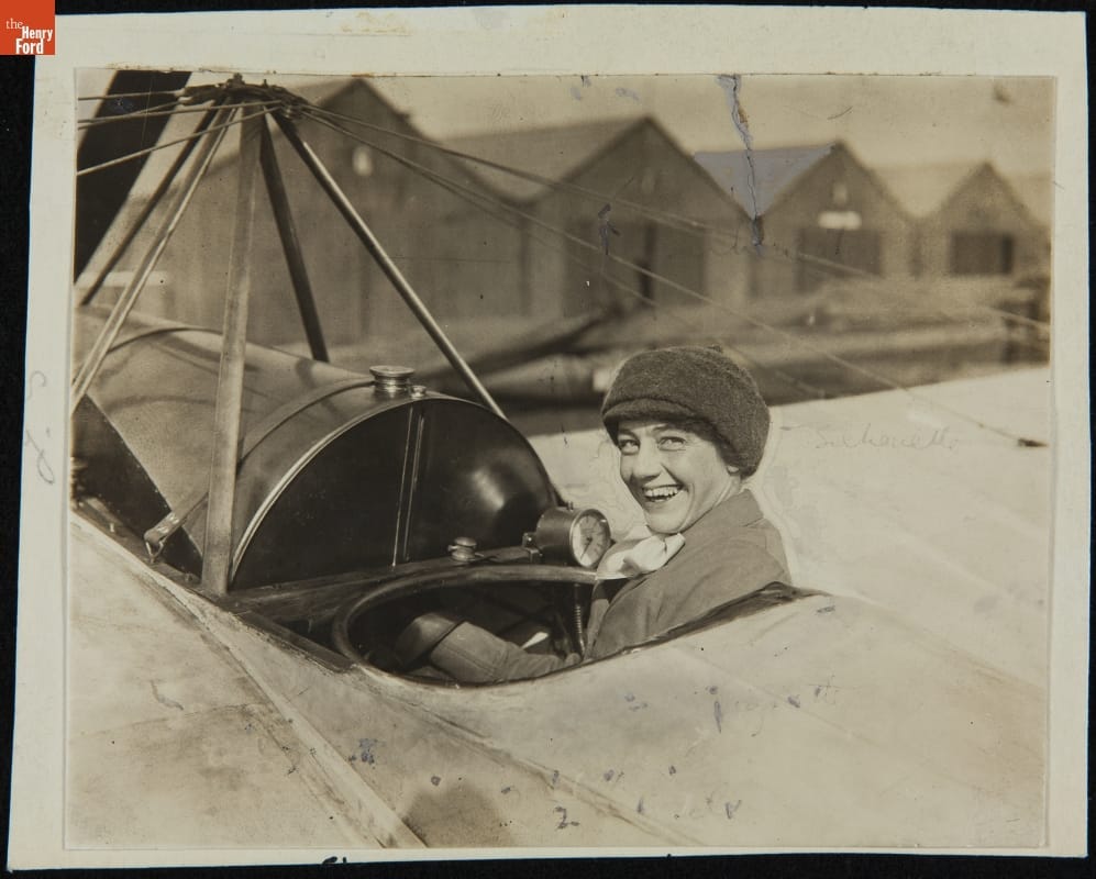Woman in an open plane cockpit smiles at the camera; a row of sheds is in the background 