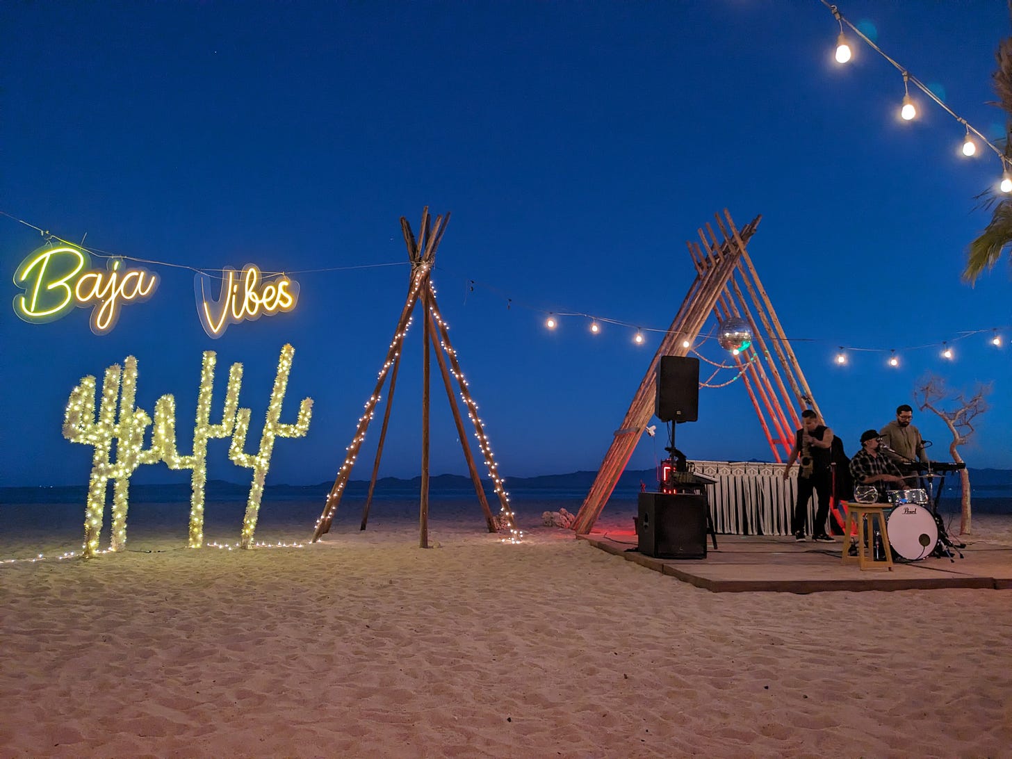 Structures set out on the beach: stage with small band, plastic cactus with twinkle lights, neon "Baja Vibes" sign