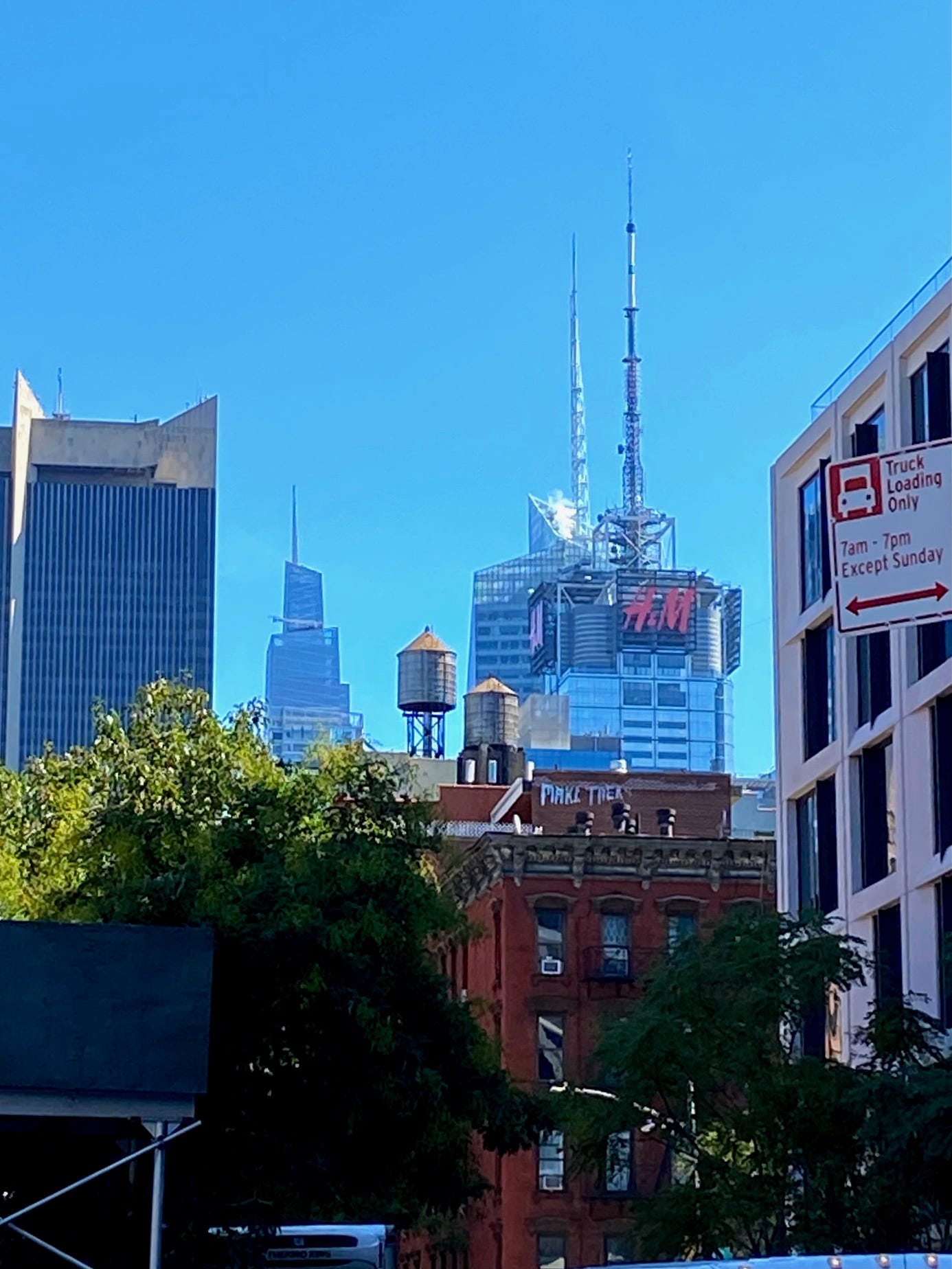 Two water towers, one on a tall pedestal, sit on adjacent roofs. In the front is a red brick building with graffiti tags. In the background is the H&M tower.