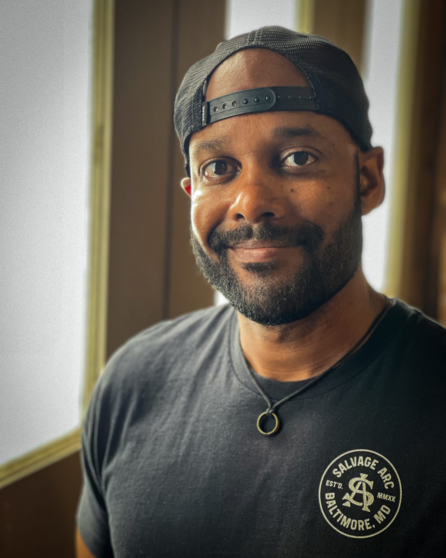 Picture of Evan Woodard. His body is angled to the viewer's left side. He wears a black baseball cap backwards, a black t-shirt with the Salvage Arc logo on it and a necklace with a silver ring on it. He has a slight smile, dark brown eyes, and a closely cropped beard and mustache. 