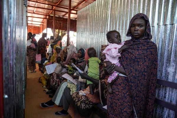 A Sudanese woman holds an infant near a line of other women with children against a wall.