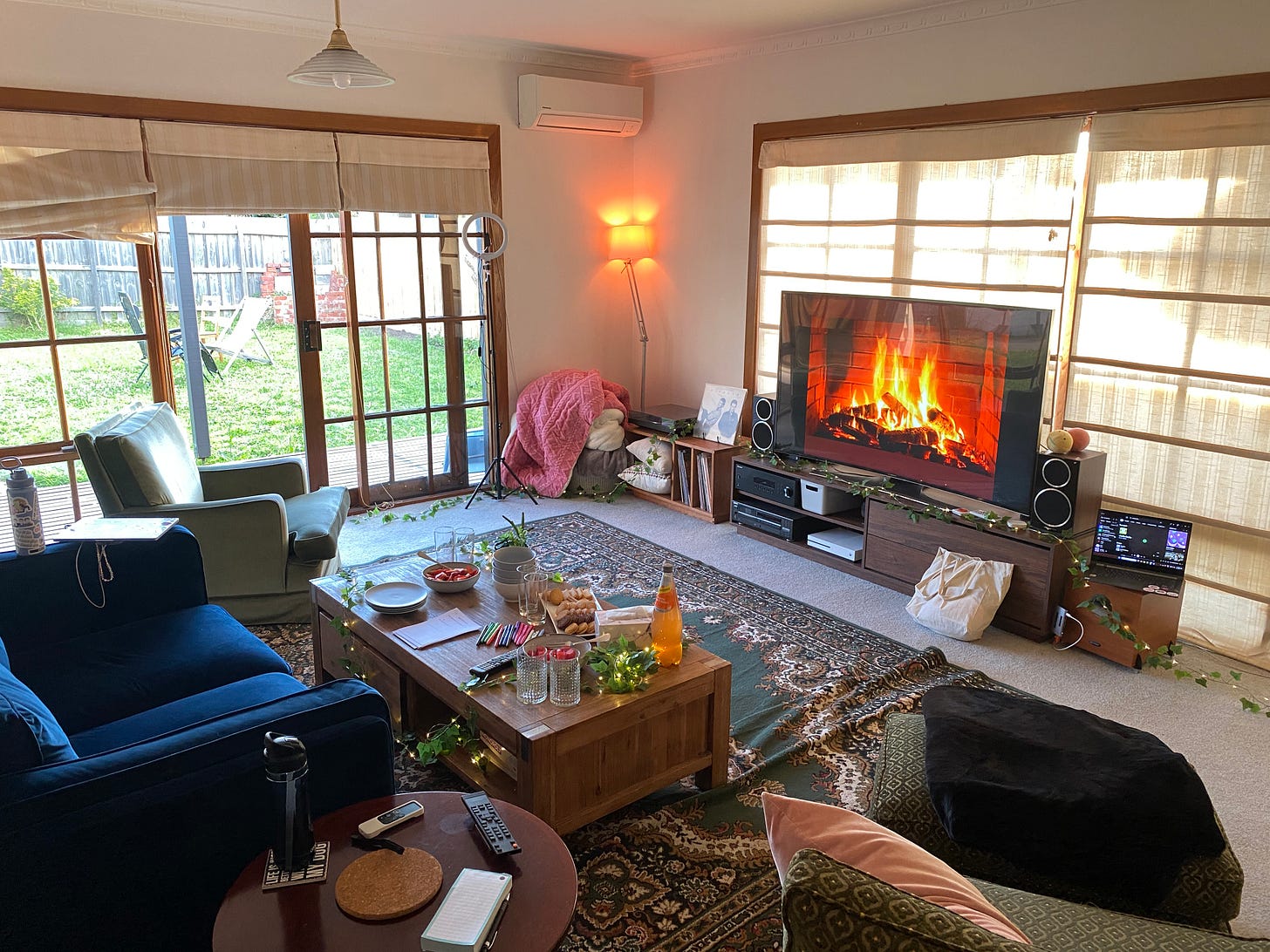A cosy living room with many comfy couches. There are fairy lights and fake leaves along the ground and an image of of a fireplace on the TV.