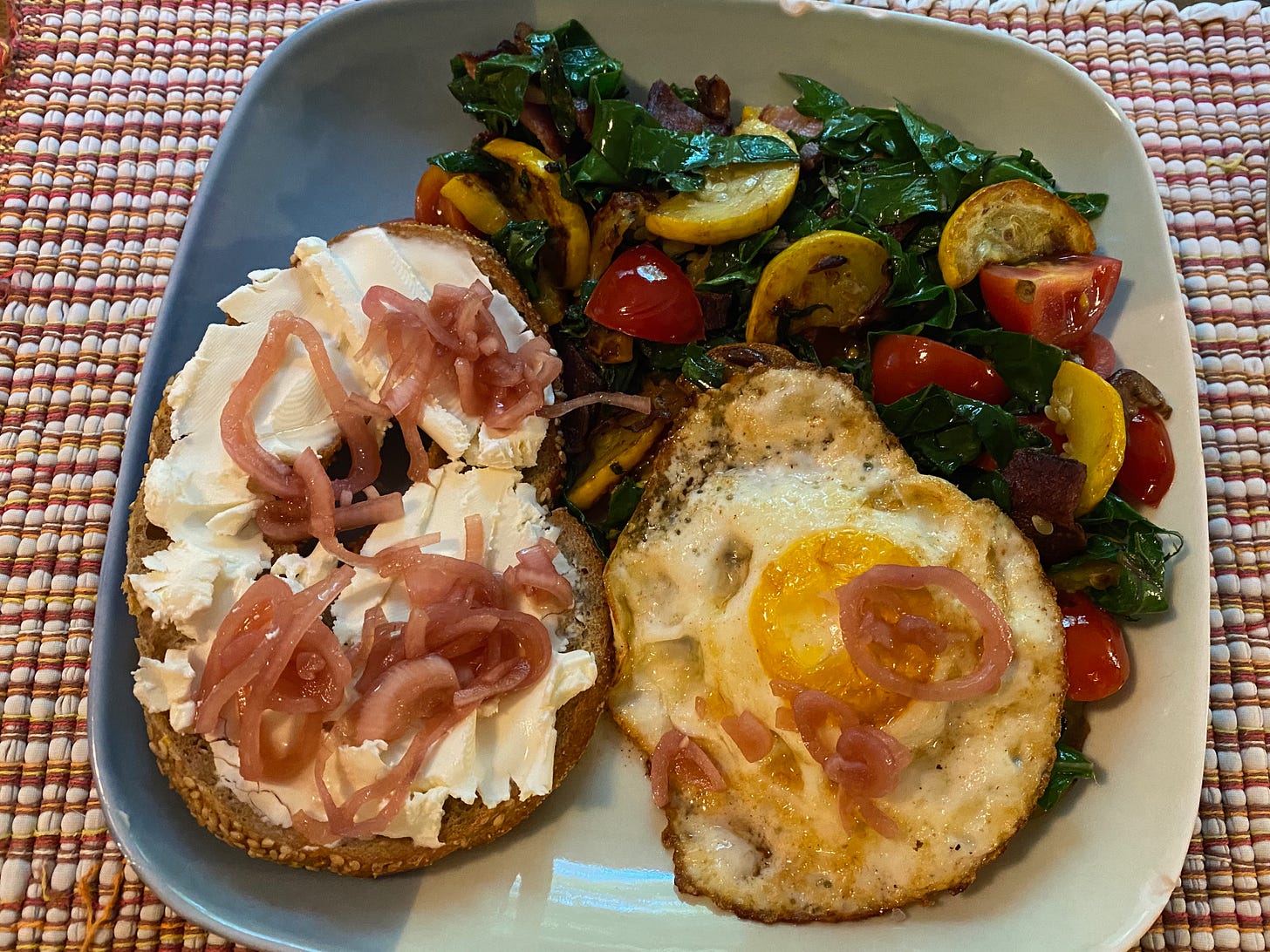 A square plate with a bagel topped with cream cheese and pickled onions, a fried egg, and a pile of sautéd kale with summer squash and chopped cherry tomatoes.