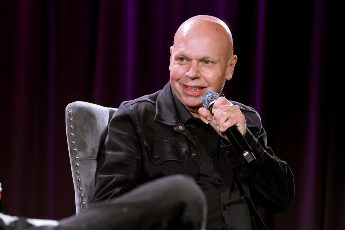 Matt Pinfield in a black jacket speaking into a microphone while he sits on a chair on a stage