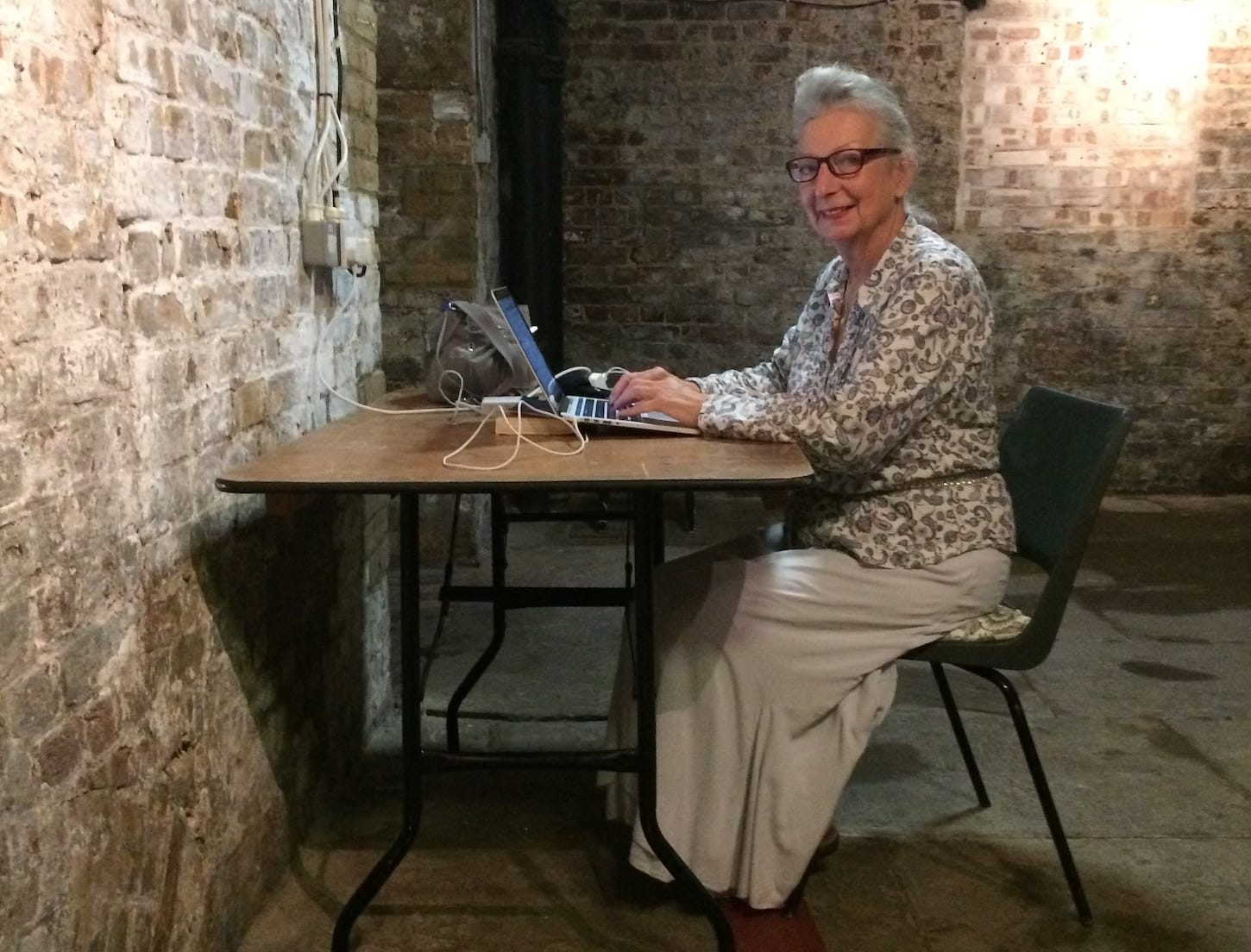 Middle aged lady wearing glasses, a blouse and a long skirt, in the crypt of a church sitting at a desk looking into the camera while typing on a laptop with old grey brown brick walls surrounding her and a flagstone floor.
