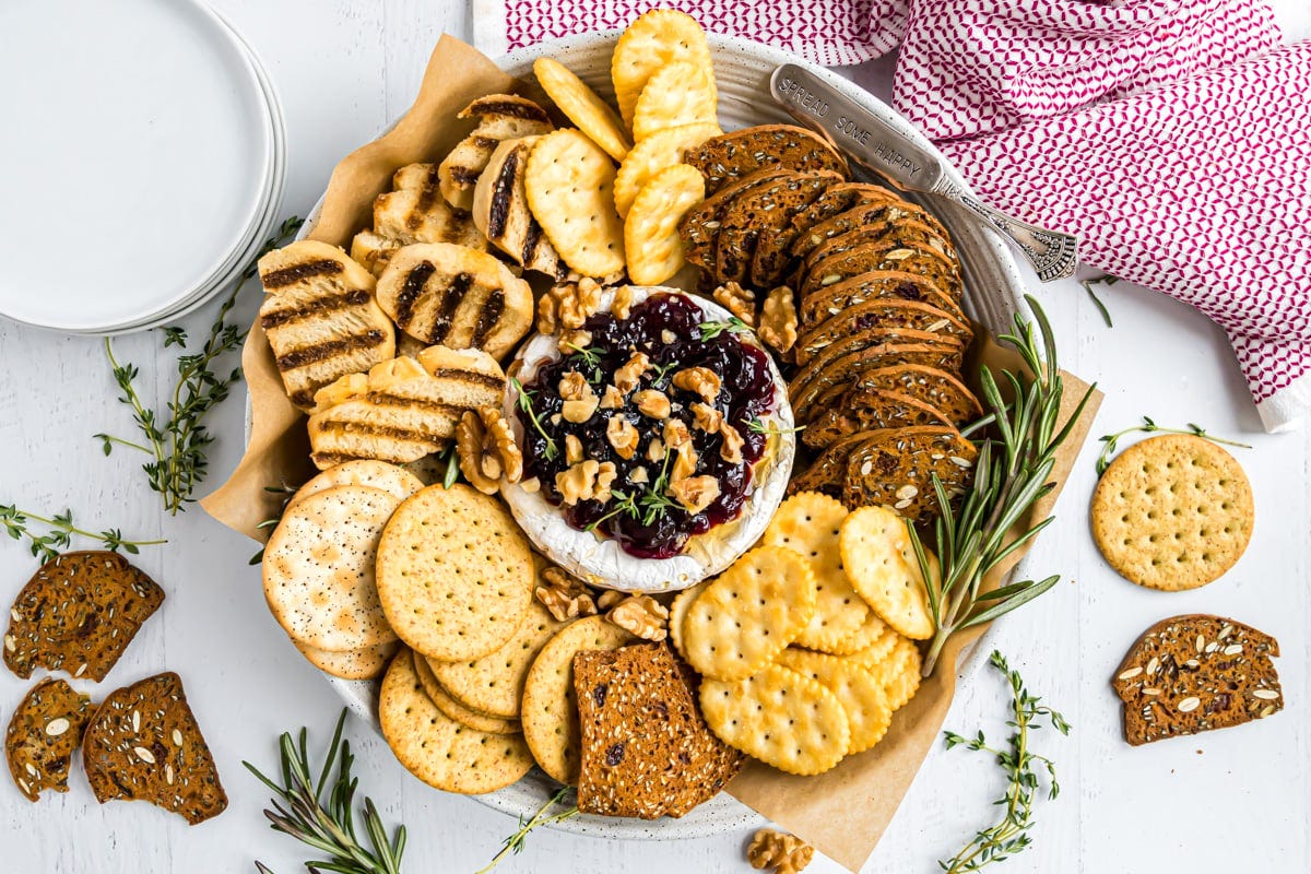 A wheel of brie on a serving platter surrounded by crackers and baguette slices.