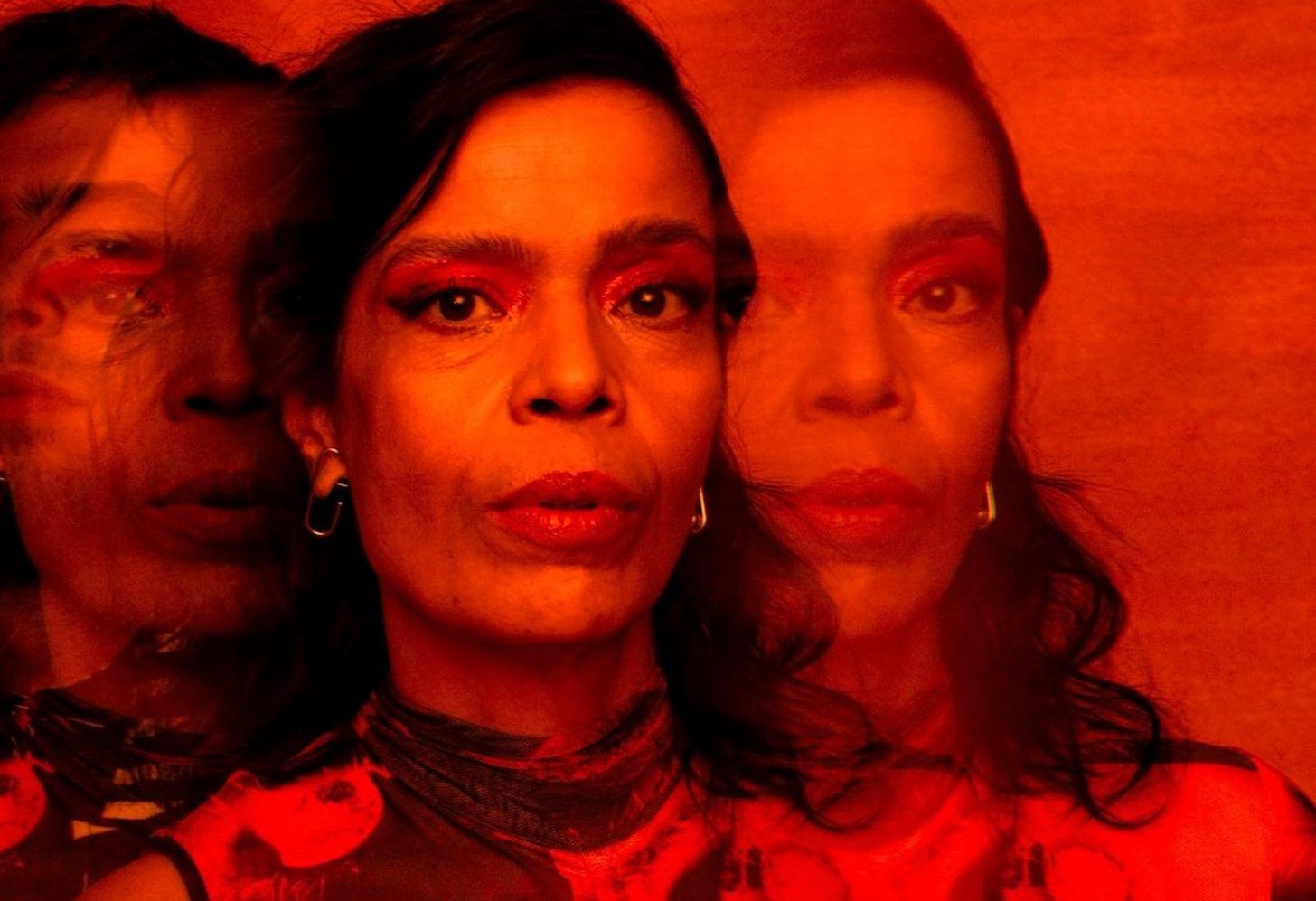 Imagem toda vermelha com uma mulher olhando para a câmera em close, sua imagem se replica como sombra nas duas laterais