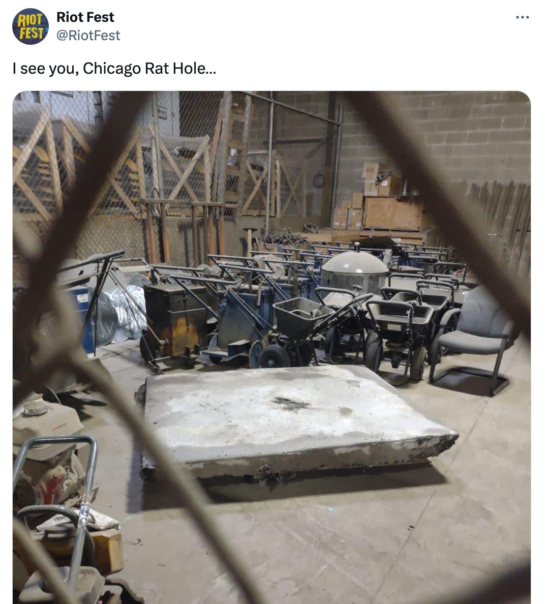 A tweet from @RiotFest that reads "I see you, Chicago Rat Hole..." and features an image taken through a chain-link fence. There's an area with a bunch of yard equipment and old chairs and stuff, and the rat hole slab is just sitting on the ground nearby.