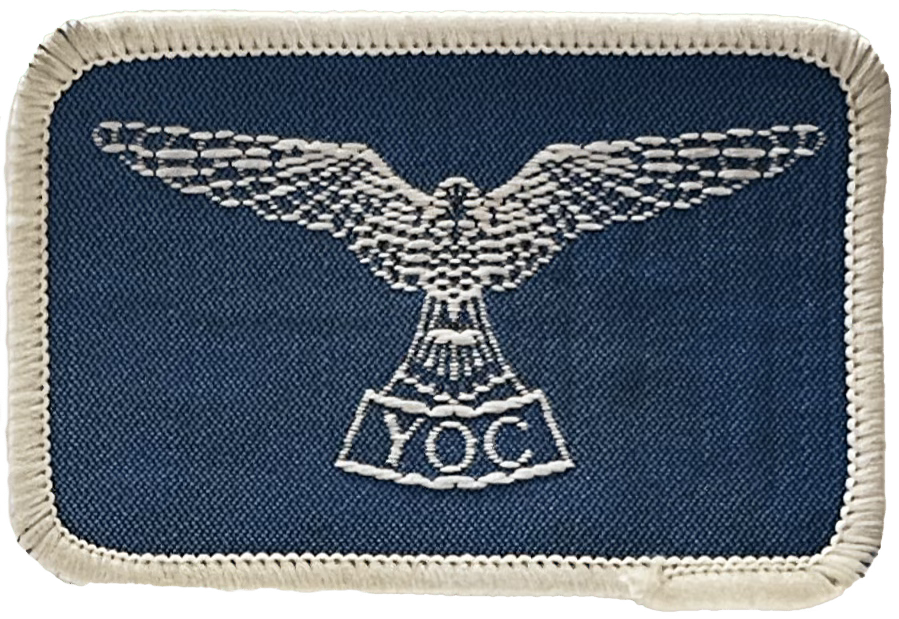 A rectangular blue cloth badge with a bird, wings outstretch embroidered in white with the letters Y.O.C underneath
