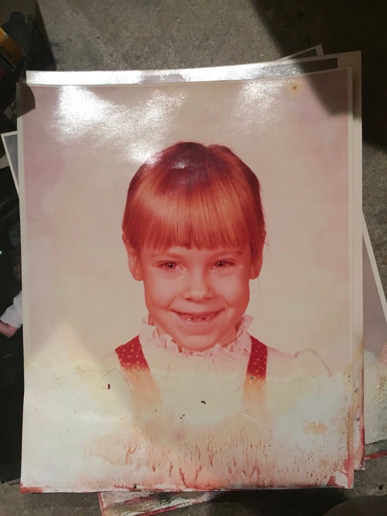 Little girl with blonde hair, missing two teeth, smiling. She wears a white blouse and a red pinafore.