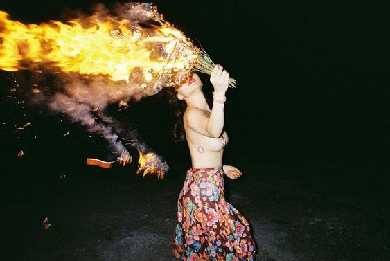 Outside in the pitch black a topless woman in a long floral skirt runs with a flaming bouquet. She holds it in front of her face so all you can see is her red lips. 