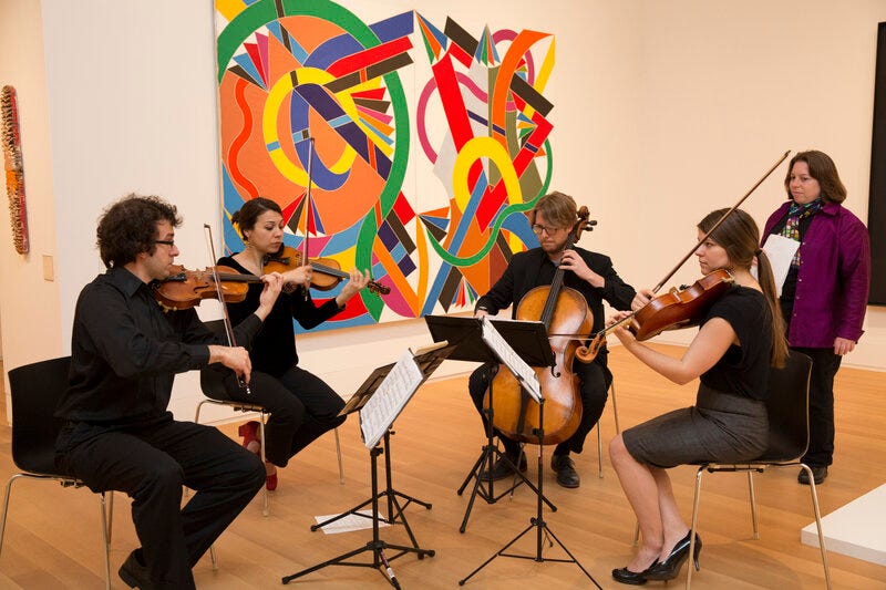 Four seated people playing string instruments in a gallery space. A colorful artwork hangs on the wall behind them. At right, a standing woman looks on. 