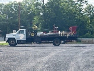 A large farm truck with a greenhouse and horse drawn tillage equipment in a parking lot