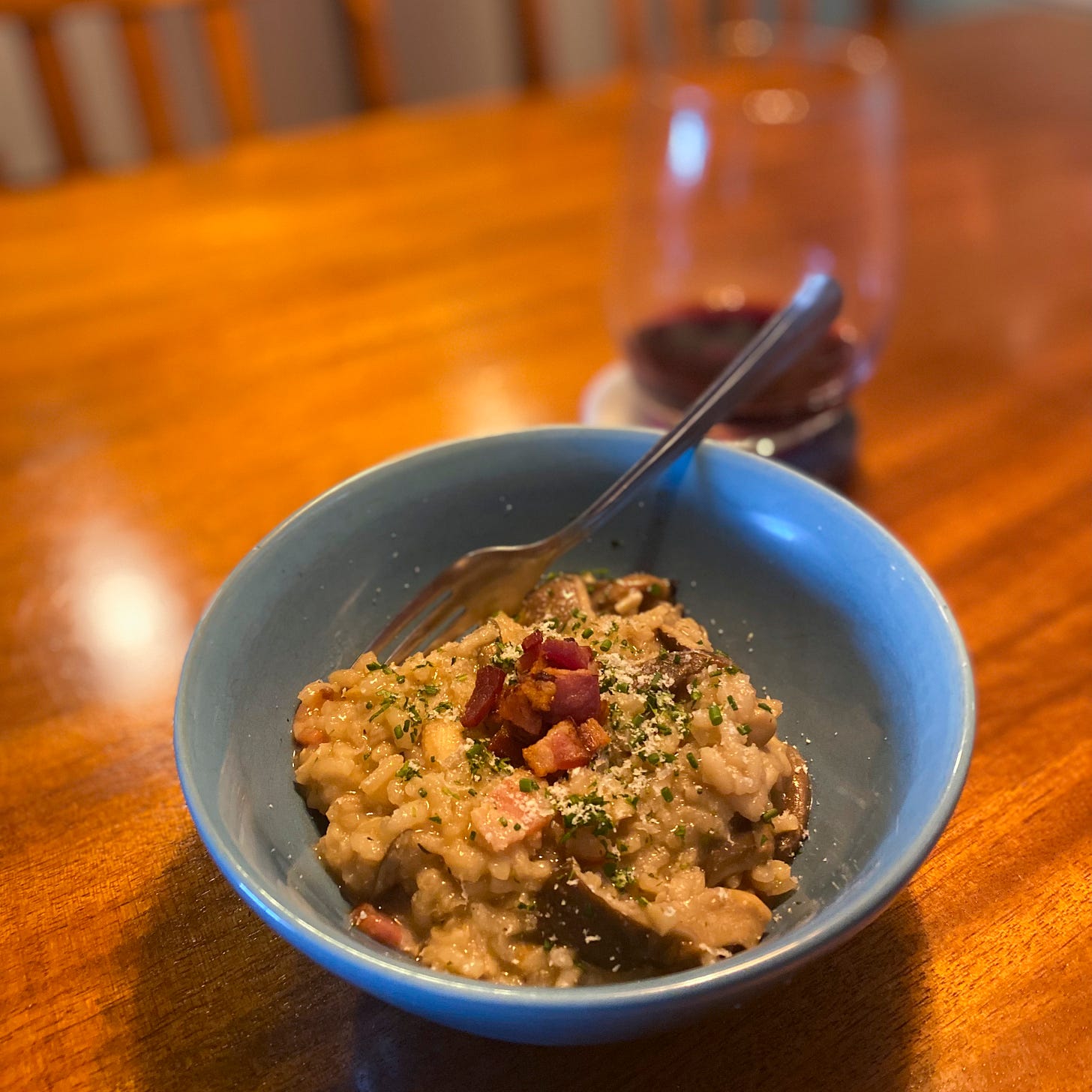 A blue bowl of the risotto, garnished with a small pile of bacon pieces, parsley, and parmesan. A glass of red wine is on the coaster behind it, and a fork rests at the side of the bowl.