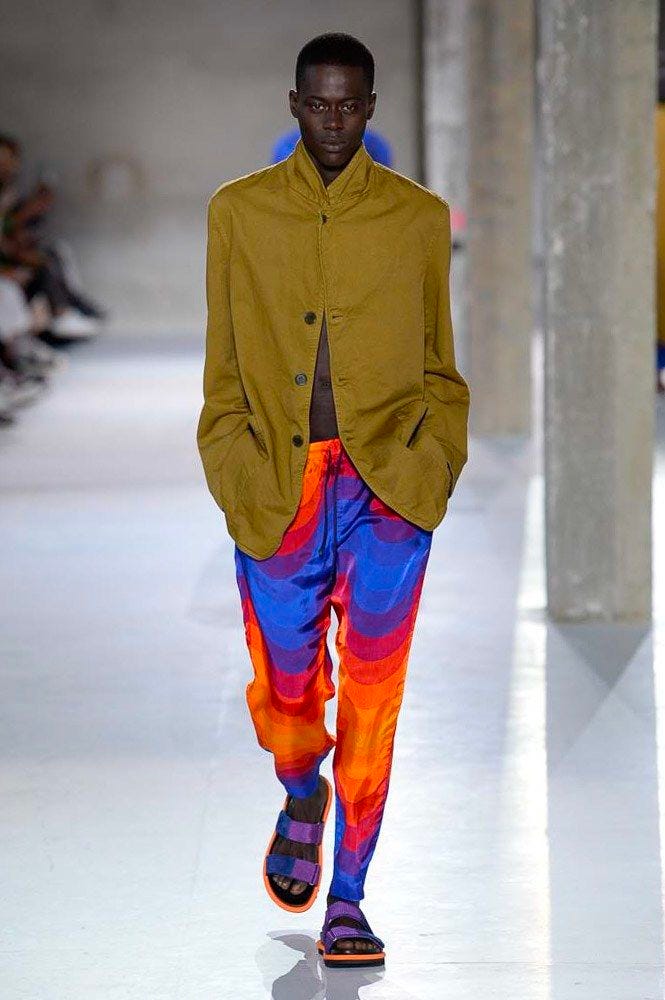 Black male model on the catwalk wearing a buttoned-up khaki style jacket and bright silk red-orange-blue-purple trousers in a swirly pattern.