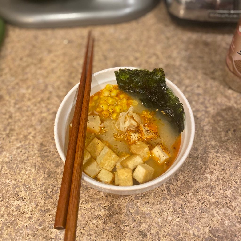 a white bowl of ramen with cubes of fried tofu, corn, and 2 sheets of roasted seaweed. The top is garnished with chili oil and sesame seeds, and a set of wooden chopsticks rest on the left edge of the bowl.