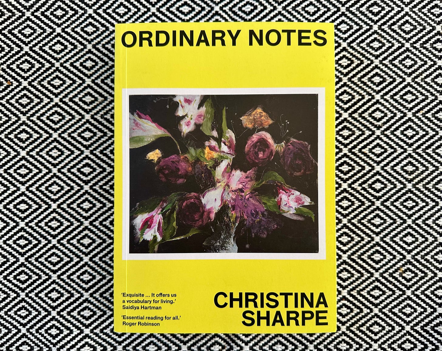 Picture of a book titled "Ordinary Notes", by Christina Sharpe, on a black and white diamond patterned cloth. The cover is a vibrant yellow, with the title and author appearing above and below an abstract untitled painting of flowers by the artist Jennifer Packer.