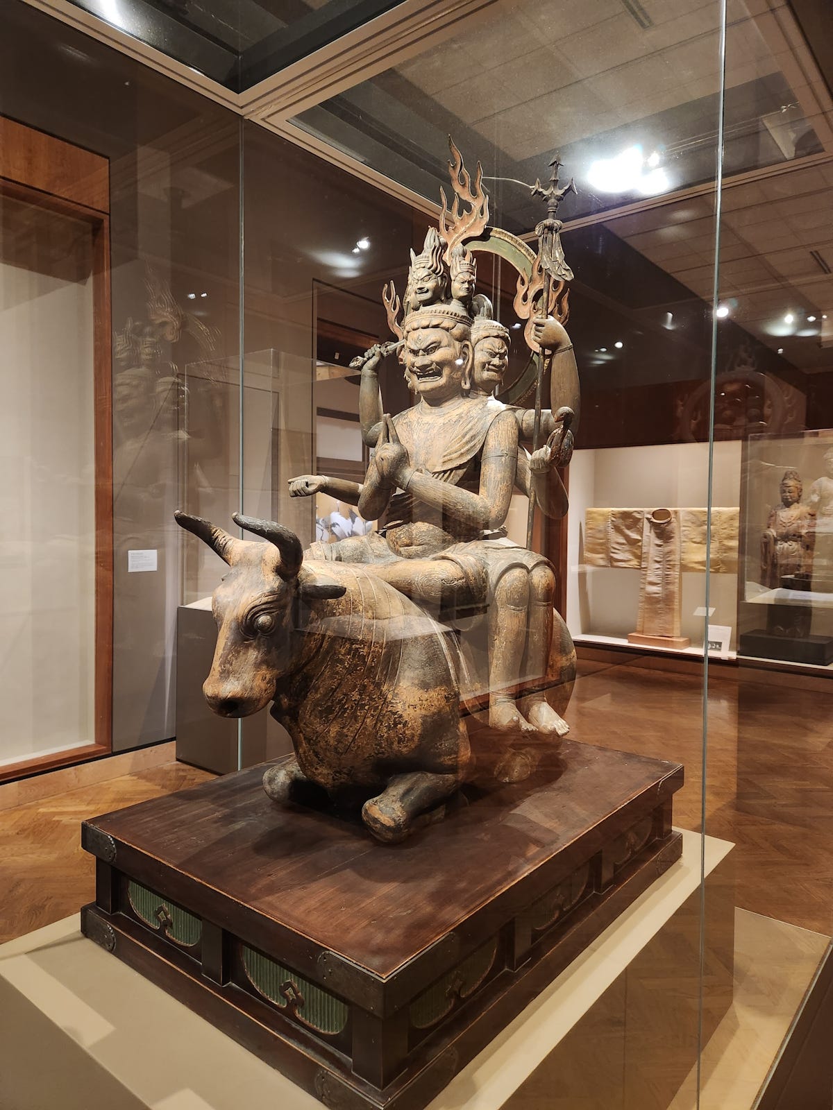 A wrathful royal divinity, carved in wood, atop a kneeling water buffalo. He has six faces, legs and arms and is riding a kneeling, pony-size water buffalo. The king’s foremost and largest face bears an expression of gleeful fury, and he holds a sword in one hand, a trident in another and, in a third, a noose for capturing people and dragging them on to the right spiritual path.