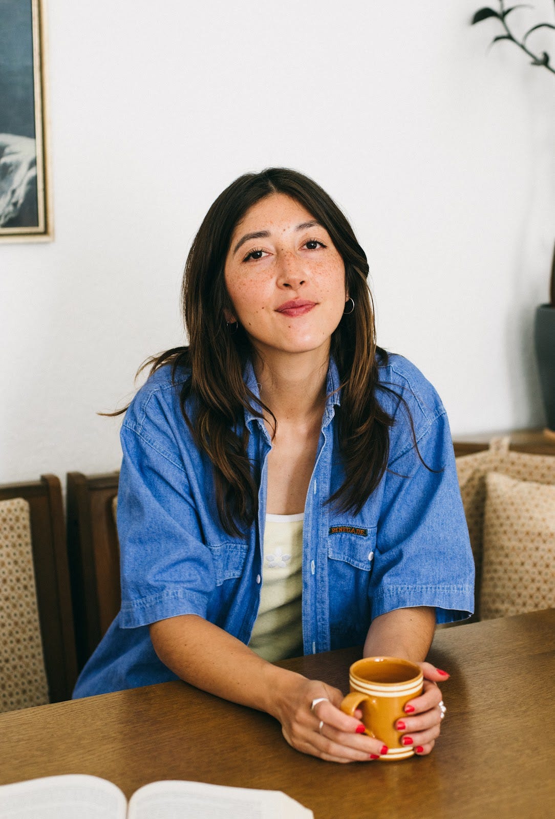 Devan wearing a blue top sat by a table holding a mug. 