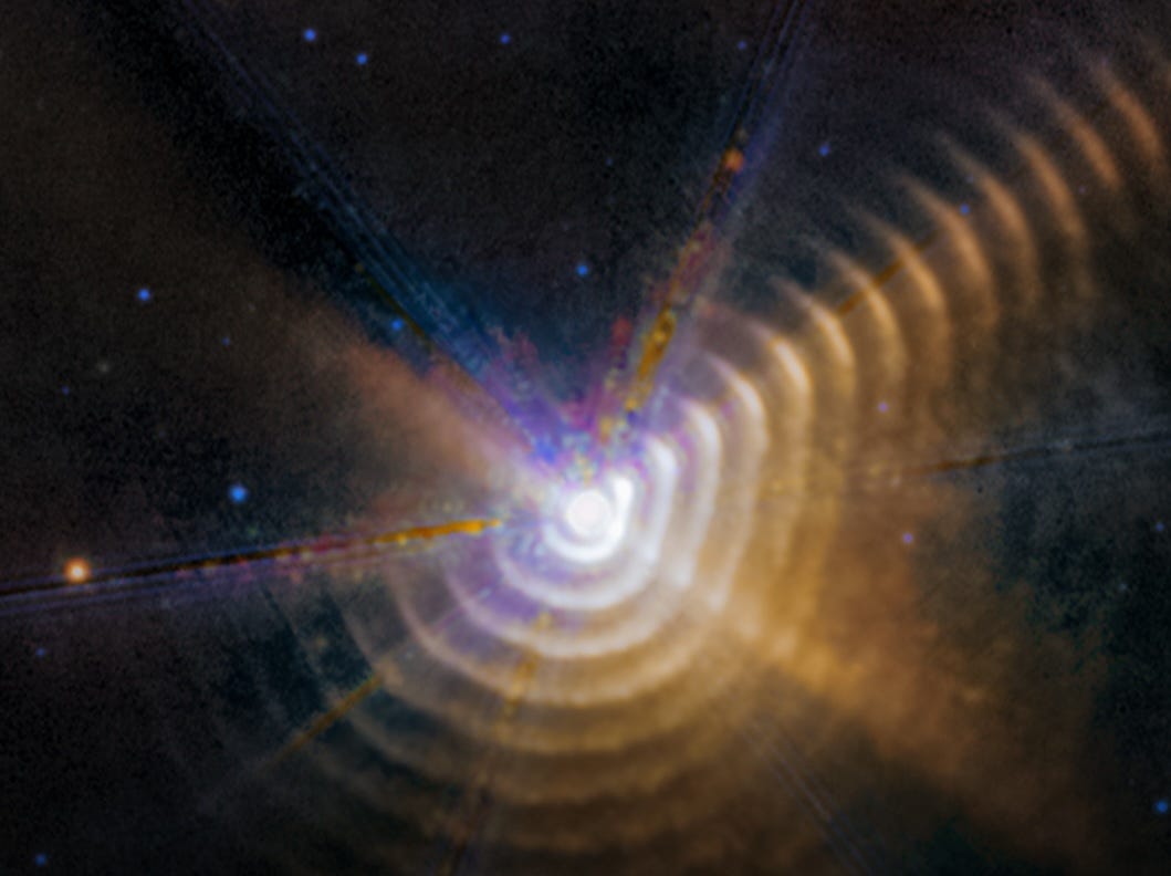 A portion of Webb’s 2023 observation of Wolf-Rayet 140. A bright white hexagon is toward the bottom left-center. This is the two stars. Blue diffraction spikes point diagonally toward 8, 11, and 1 o’clock. Surrounding the central light are a series of regularly spaced rings. A segment of each of the rings at around 2 o’clock appears brighter. These bright patches form a line that travels to the upper right. A few blue background dots are on the black background of space.