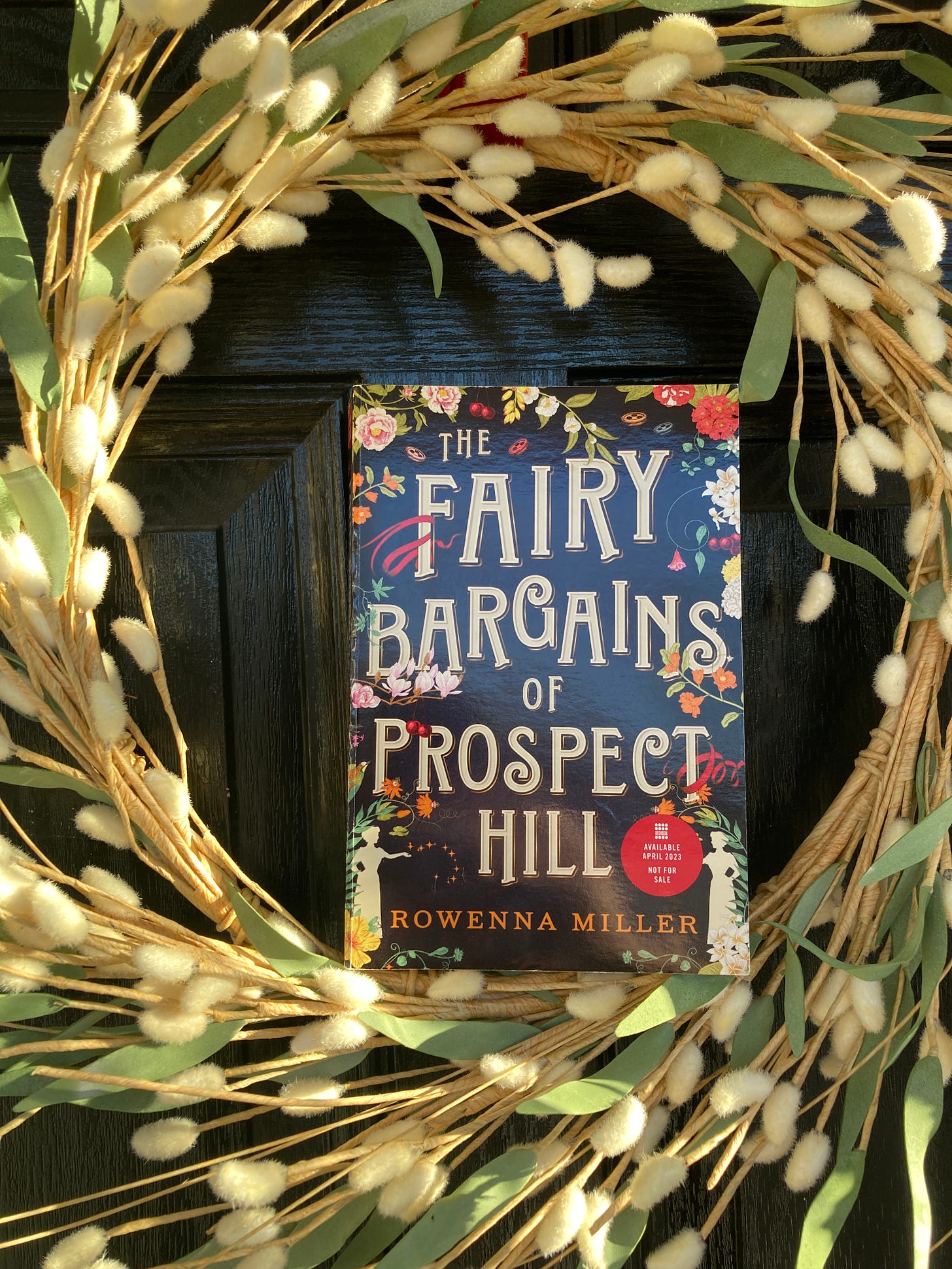 Book, THE FAIRY BARGAINS OF PROSPECT HILL by Rowenna Miller, set within a wreath of white and green willow branches against a black door