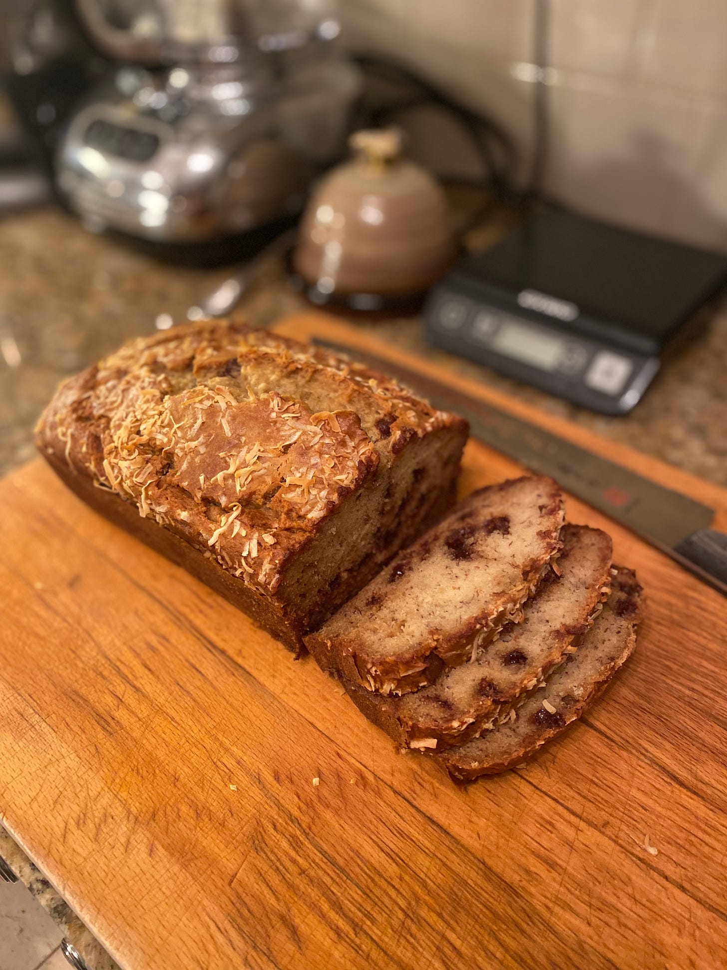 On a wood cutting board, a loaf of banana bread with a crisp-looking crust and a split across the top, baked-in coconut scattered over it. It's been sliced open so you can see the light colour inside, and the chocolate chips. Three slices tumble onto the cutting board in front, next to the bread knife.