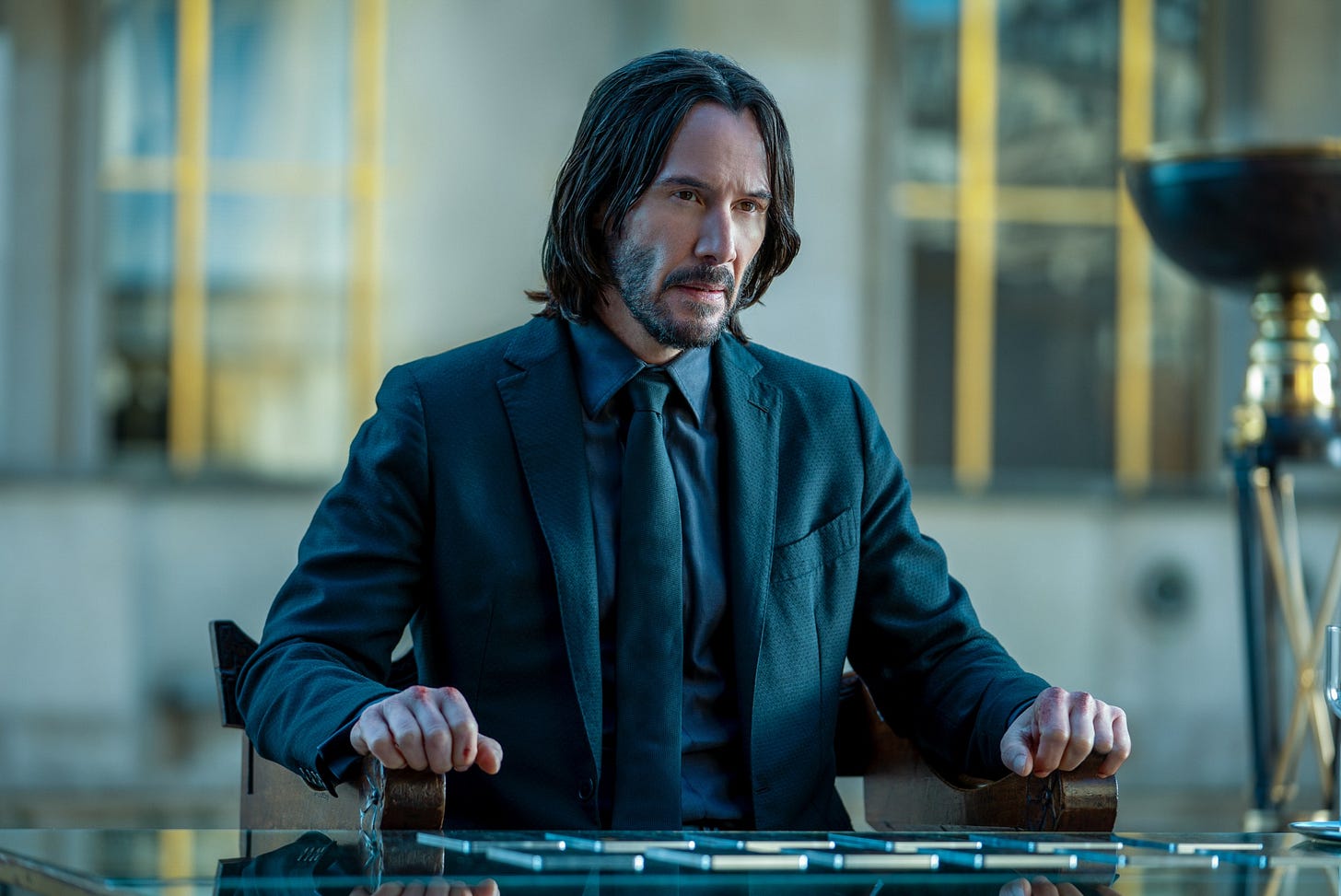 A man with long black hair, wearing a black suit with a black shirt and tie who is sitting at a table with his arms resting on both arms of a chair.