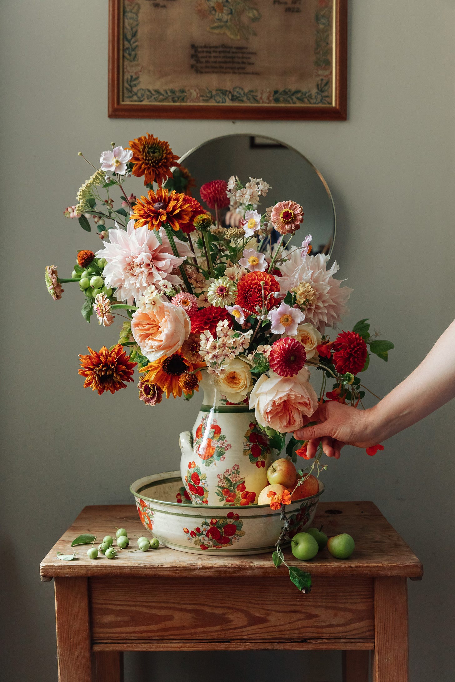 An arrangement of summer flowers of all shapes and sizes in a jug. The jug is ceramic and sat in a matching bowl on a wooden table against a wall. There is a framed embroidery sample above the flowers and a hand coming into frame on the right to adjust the flowers.