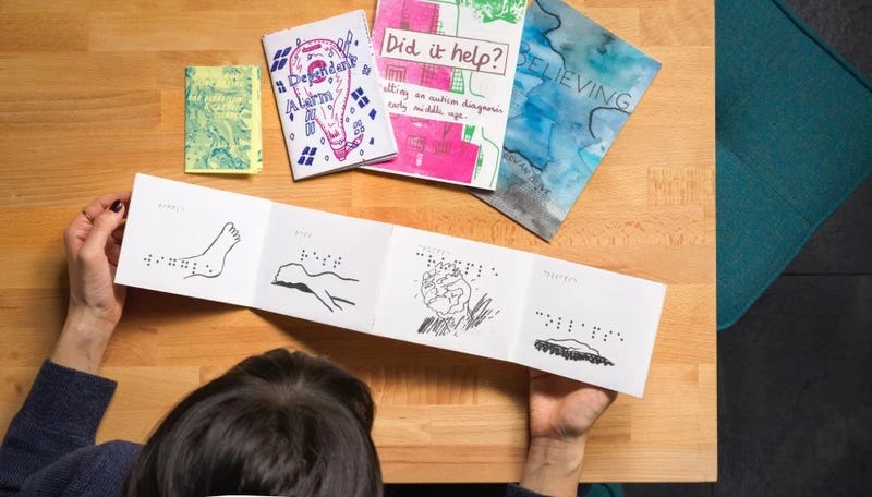 A person with dark hair, wearing a dark-colored sweater, is seated at a wooden table, holding an open accordion-style book featuring simple black ink drawings with Braille text.