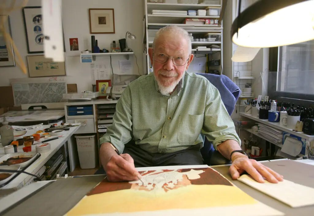 A color photo of a cartoonist sitting at his drawing table under a lamp. He has short gray hair, gray Van Dyke beard and wire-rimmed glasses. He wears a green shirt with its sleeves rolled up. 