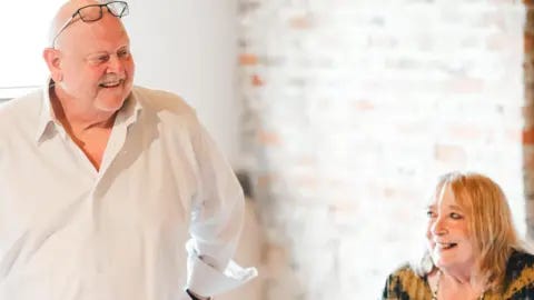 Norfolk Police  Peter Darby standing and smiling, wearing a white shirt and with spectacles on top of his head. Kathleen sits beside him, smiling at him, wearing a black and mustard patterned top. 