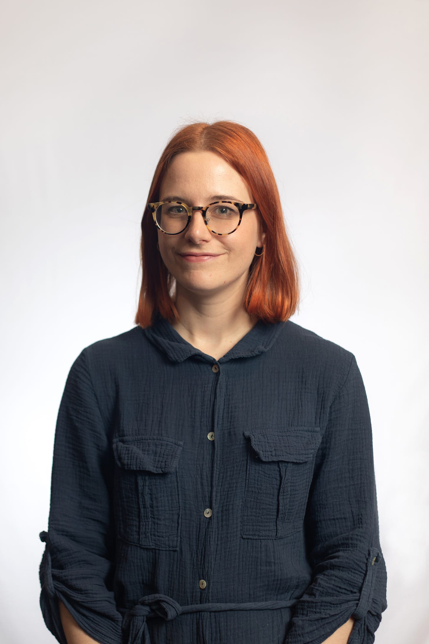 Victoria has fair skin and straight copper-red hair. She's wearing large round tortoise glasses and a petrol blue linen shirt dress. She's smiling with her mouth closed and staring straight into the camera.