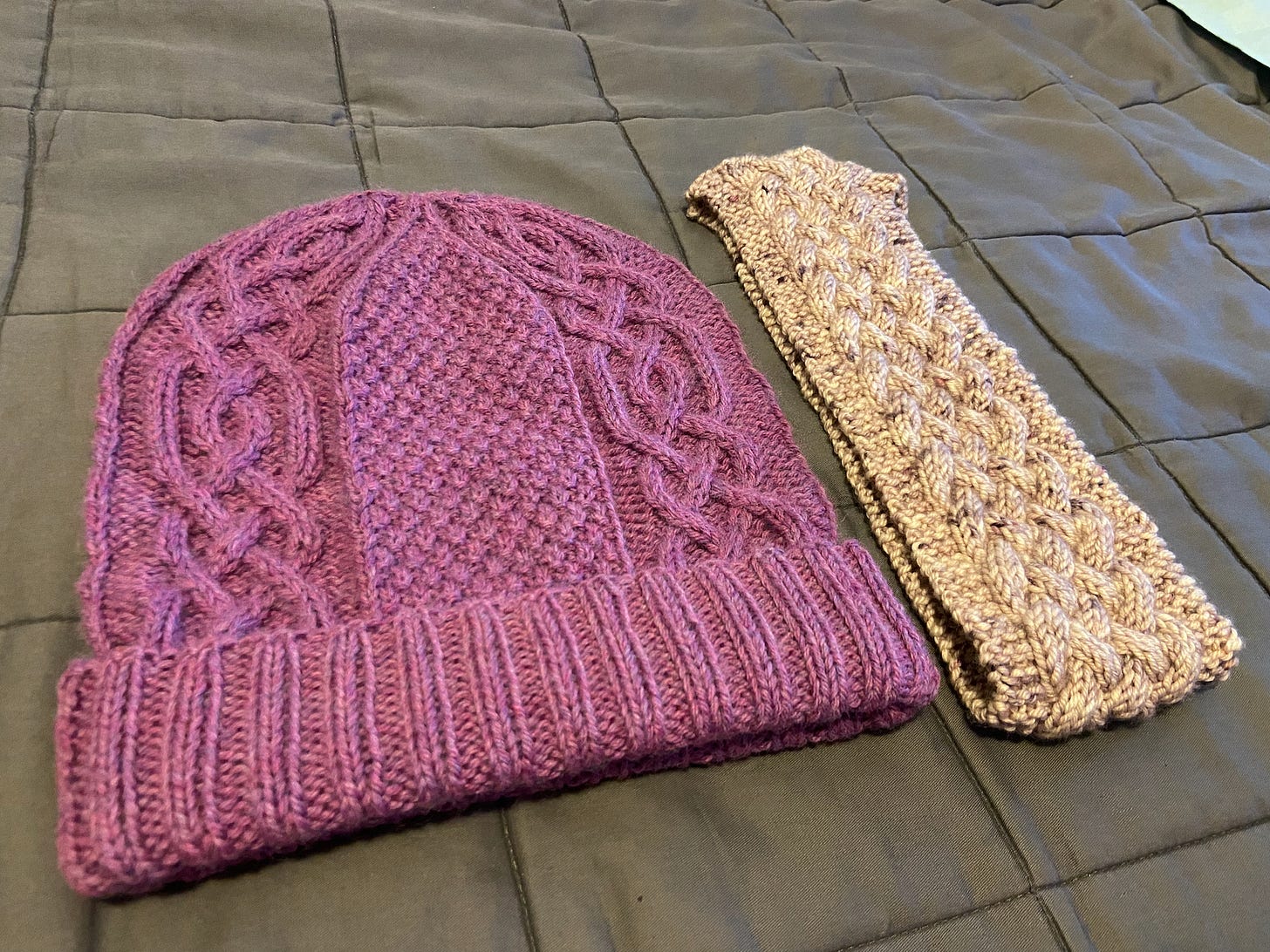 The Norway Pine Hat in purple (left) and the Knotwork Cabled Headband in a light lilac (right). They're both positioned on top of a comforter.