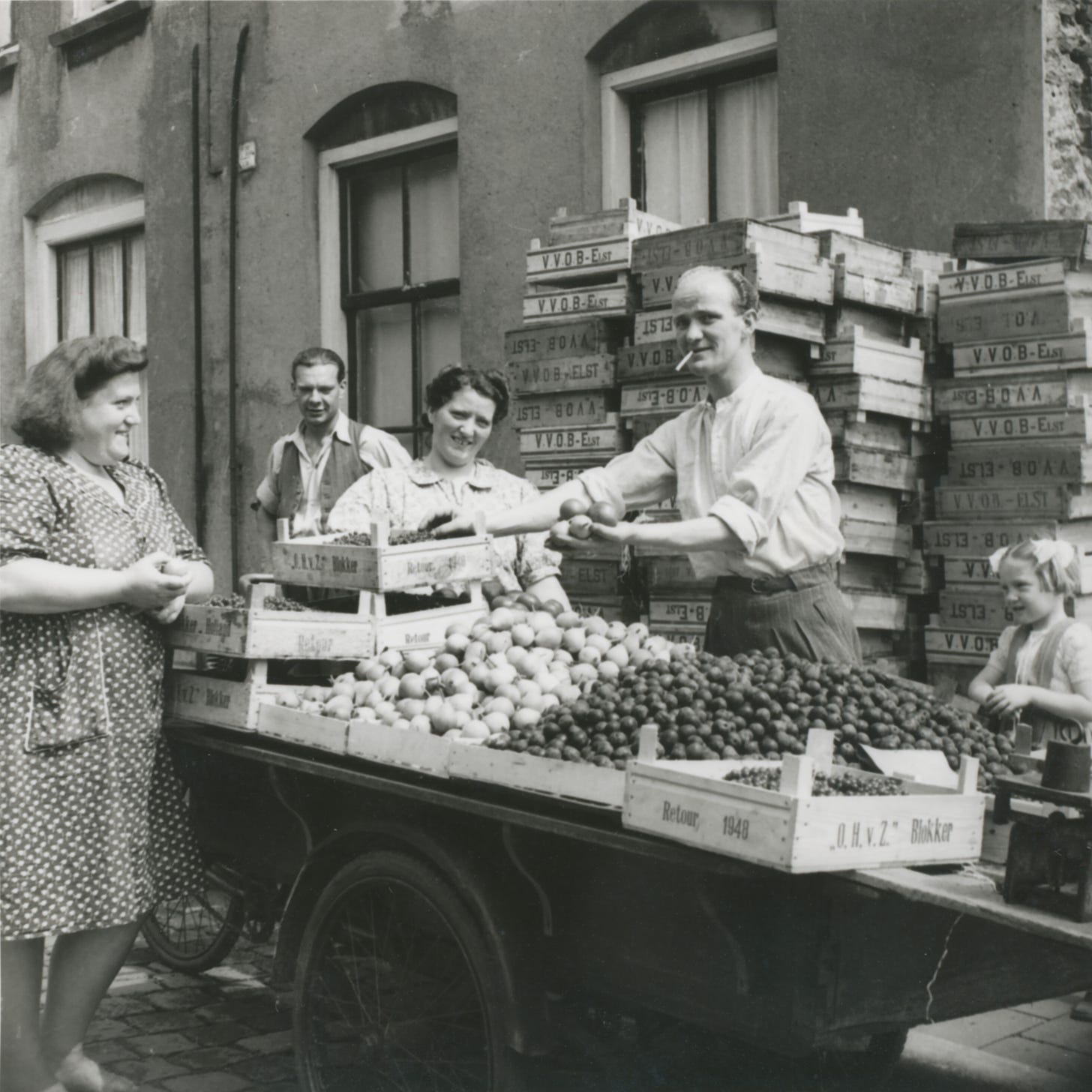 An old black and white photo showing a man stood behind a large cargobike. The bike is covered in boxes of vegatables which the man is selling. A women is stood in front of the bike purchasing some potatoes 