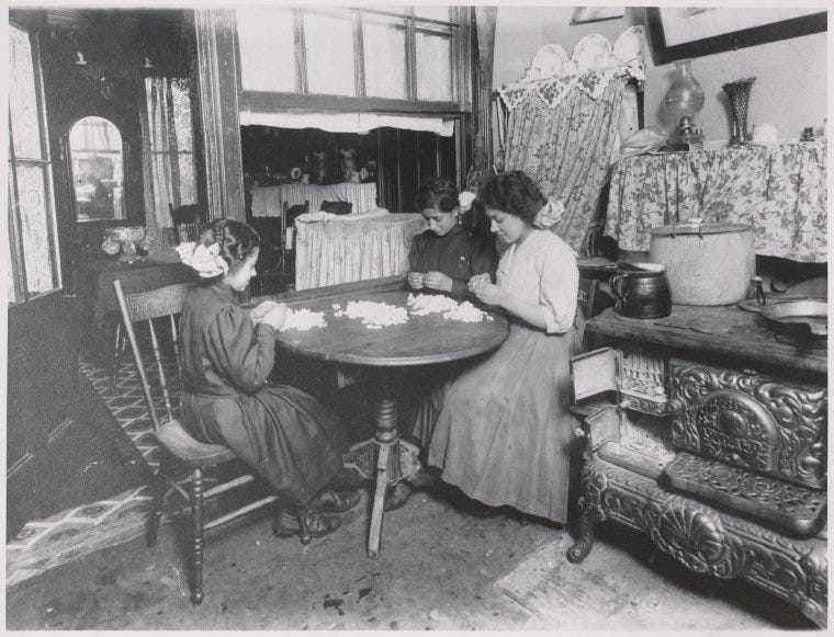 An antique photo of two women and a girl making cloth flower at a table in a small kitchen. 