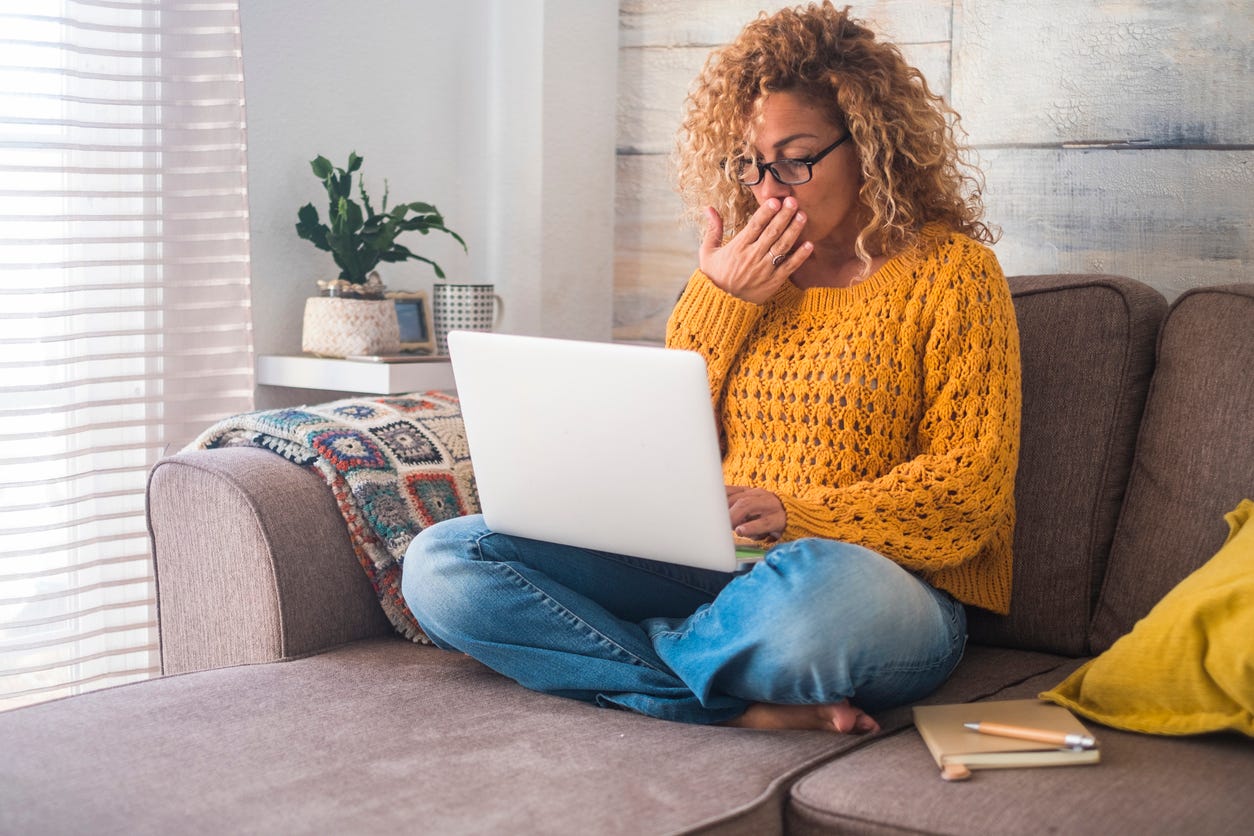 Young woman sitting on sofa with laptop. She has her hand to her mouth as if she's surprised at what she's seeing.