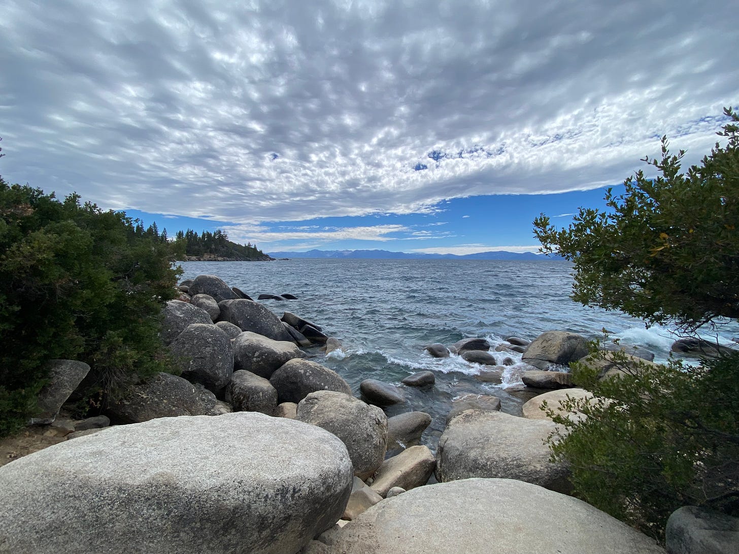 A photo of Lake Tahoe with rocks and trees on the shoreline, and a dramatic blue sky with gray and white clouds bunching up overhead.