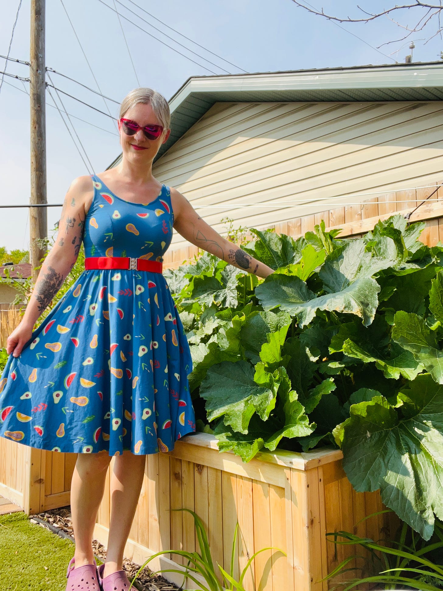 Photo of Jen wearing a blue dress with colourful veggie pattern standing next to the large zucchini plants.