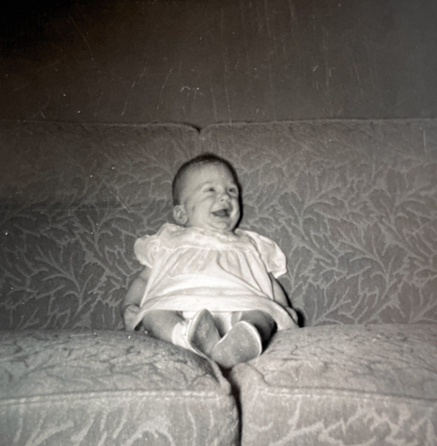 A four-month old baby, sitting alone on a couch laughing. 