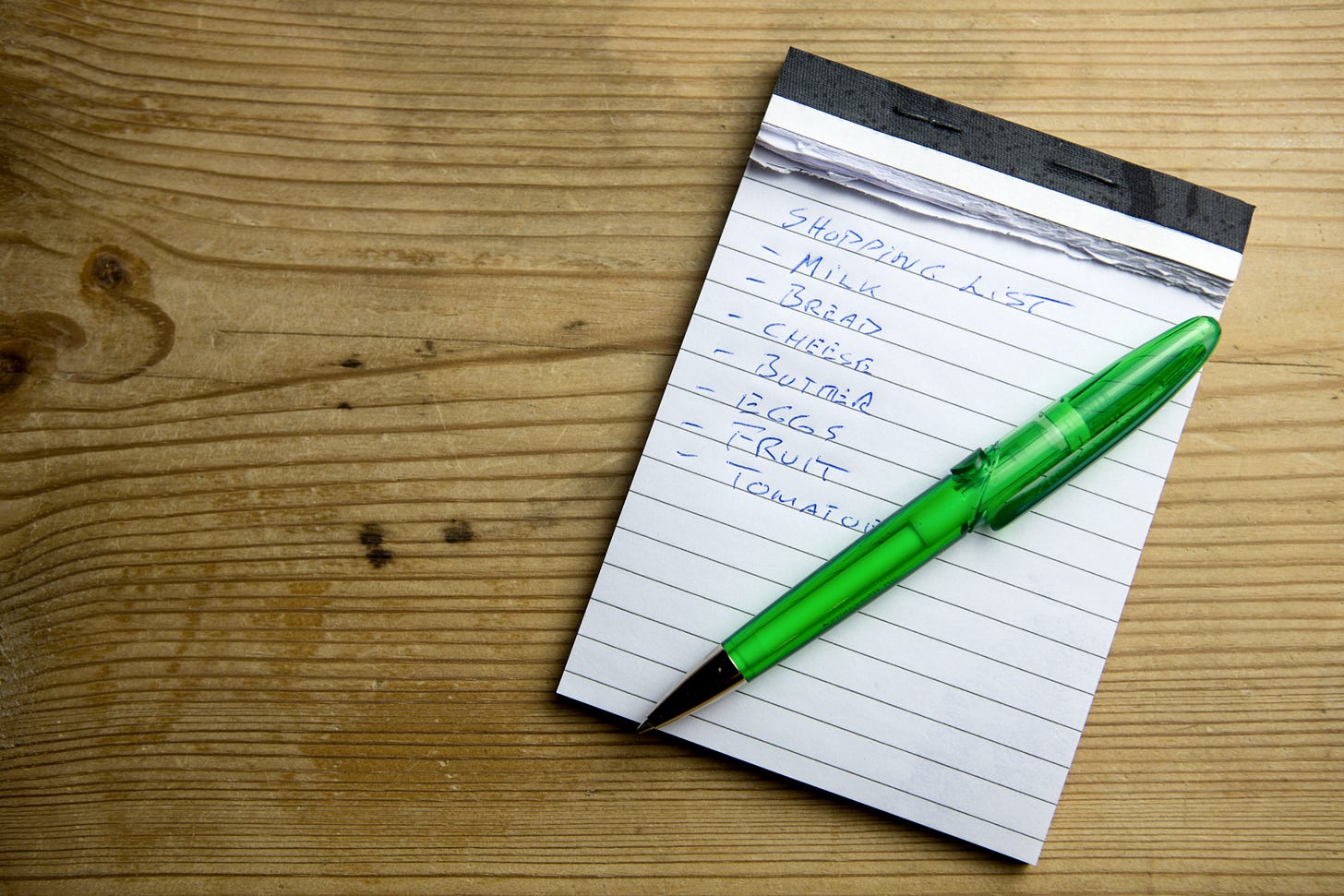 A white notepad with black lines sits on a brown wooden table. A green pen rests on top of the notepad.