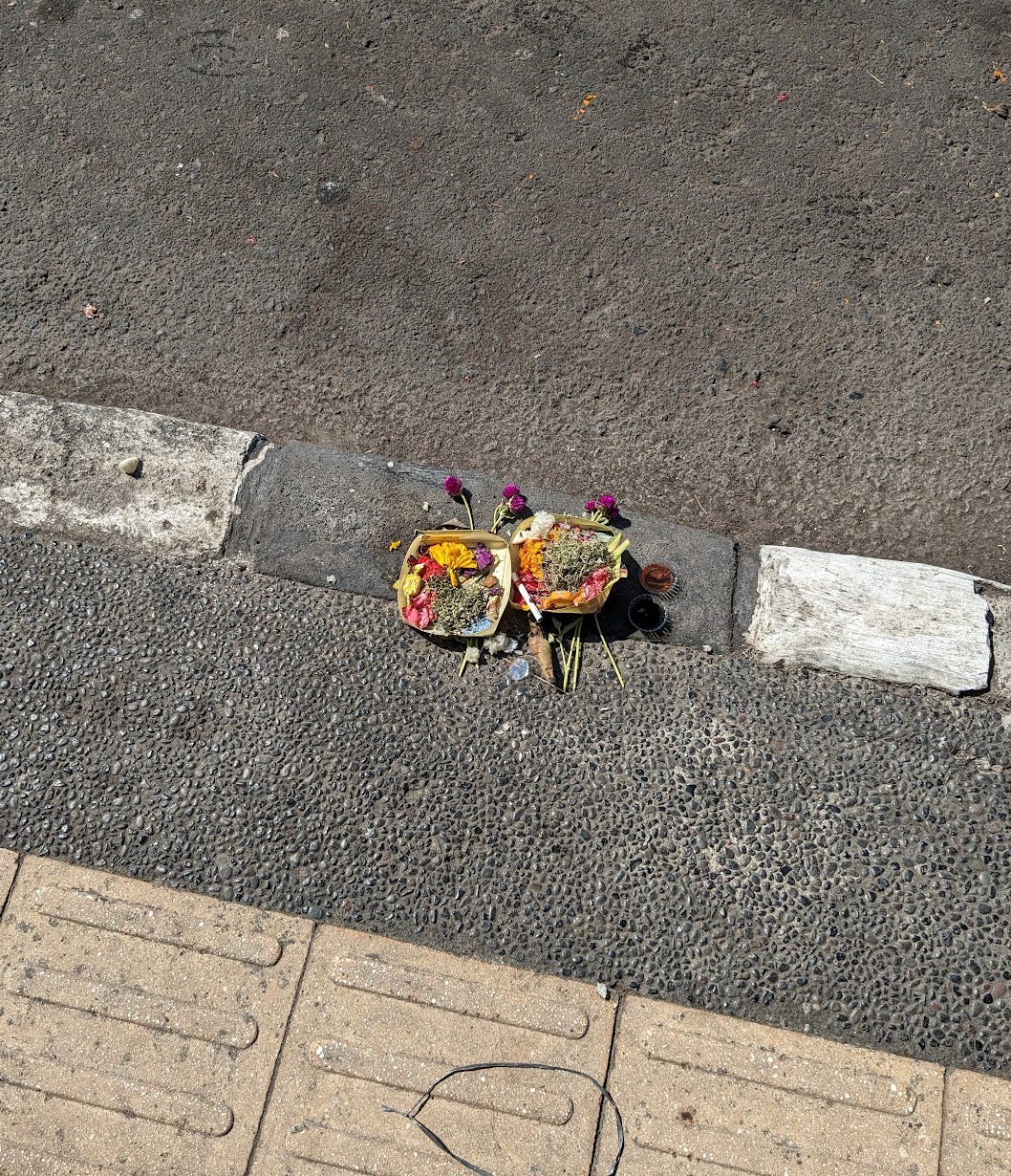 Yellow, purple, and white flowers are placed in banana leaves that were shaped into a bowl shape to hold in the offerings. The offering is placed on the edge of a sidewalk